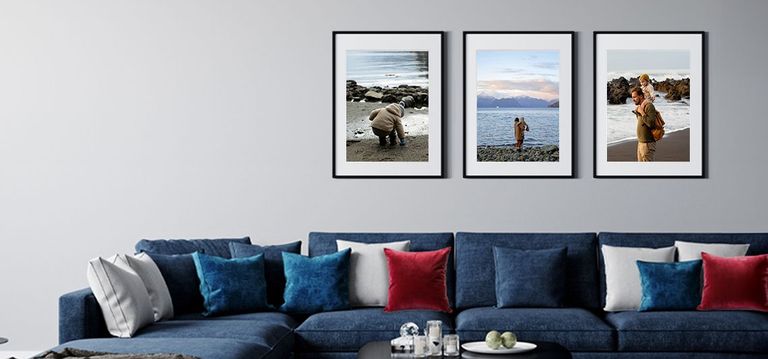 Three framed photos of man and his son walking near water framed in black frames with white mats above a blue sofa with red white and blue accent pillows